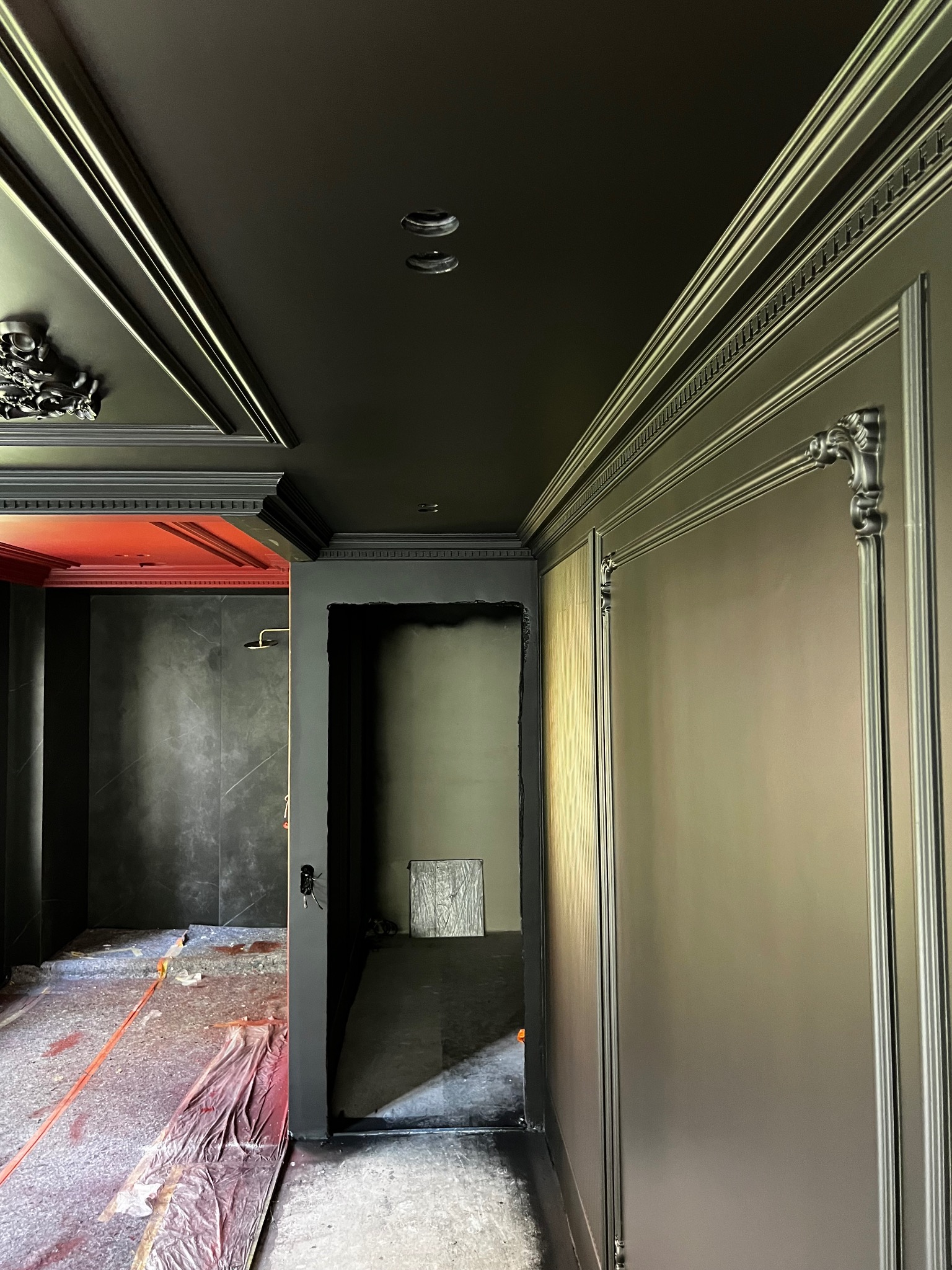 Eleganckie wnętrze w trakcie malowania: czarne ściany z dekoracyjnymi listwami, zabezpieczona podłoga, widok na łazienkę z szarą glazurą i czerwoną wnęką.