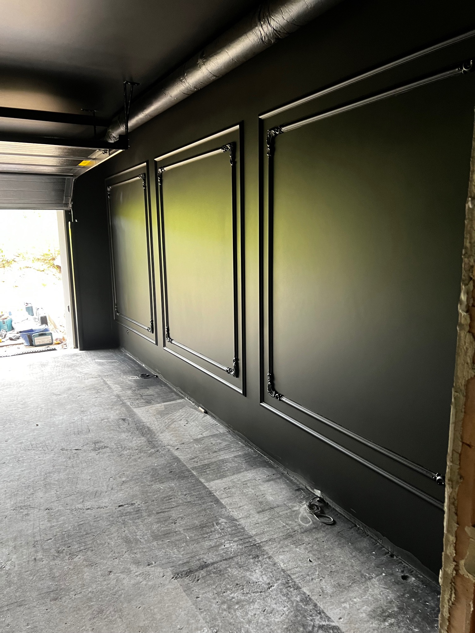 Eleganckie, czarne ściany z dekoracyjnymi ramami w nowo wykończonym wnętrzu, widok na otwartą bramę garażową i surową posadzkę.