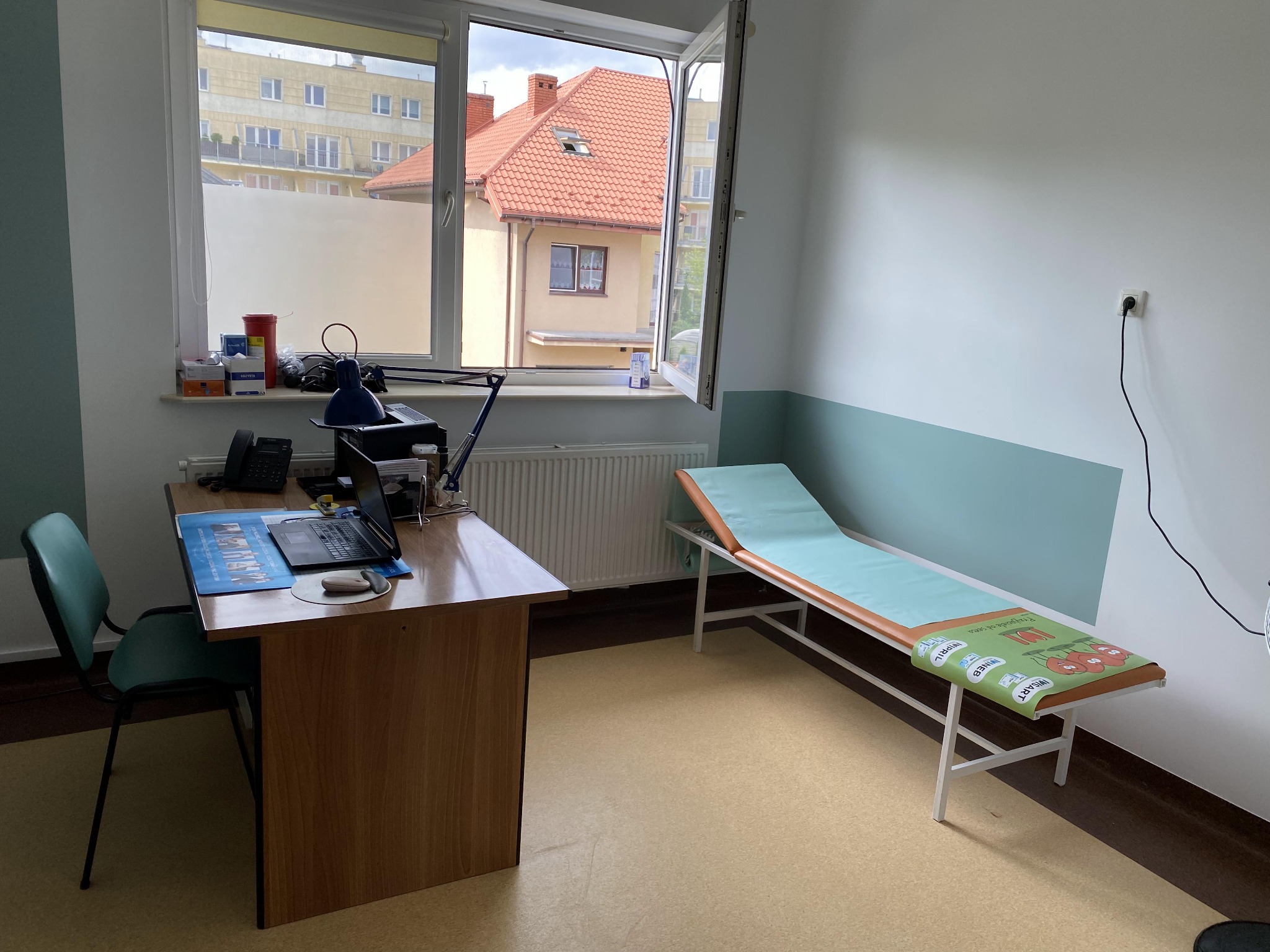 Jasne wnętrze gabinetu lekarskiego po odświeżeniu: biurko z laptopem, kozetka z zielonym podkładem, ściany pomalowane na biało z turkusowym pasem na dole.