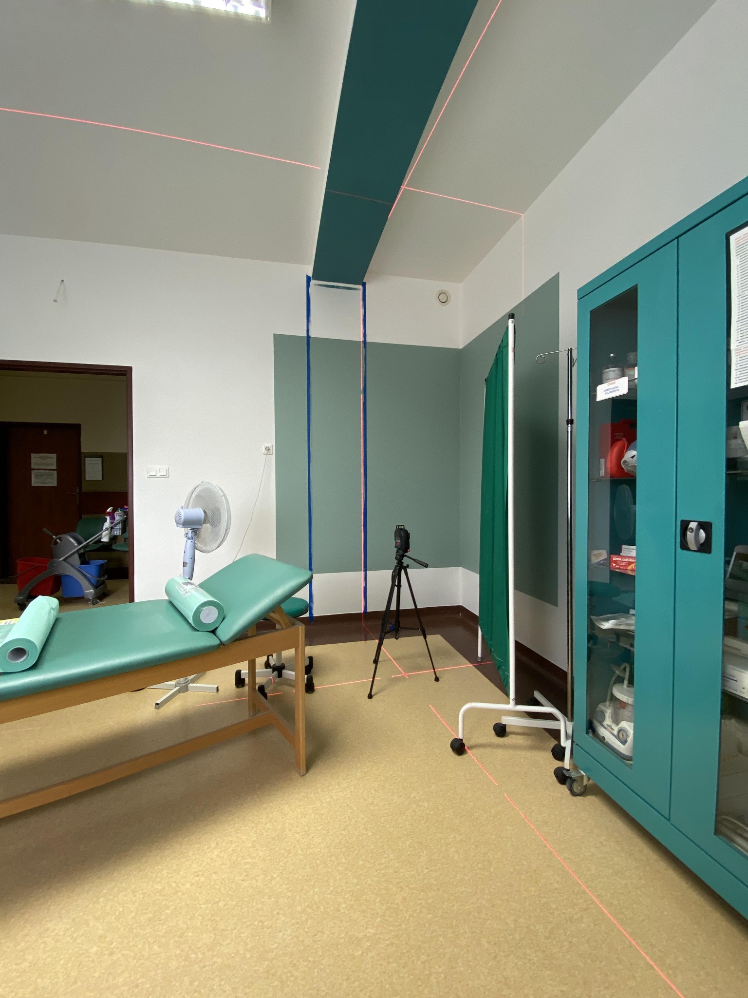 Pokój z laserowo wyznaczonymi liniami na ścianach i suficie, przygotowany do prac wykończeniowych. Widoczny sprzęt medyczny i wentylator.