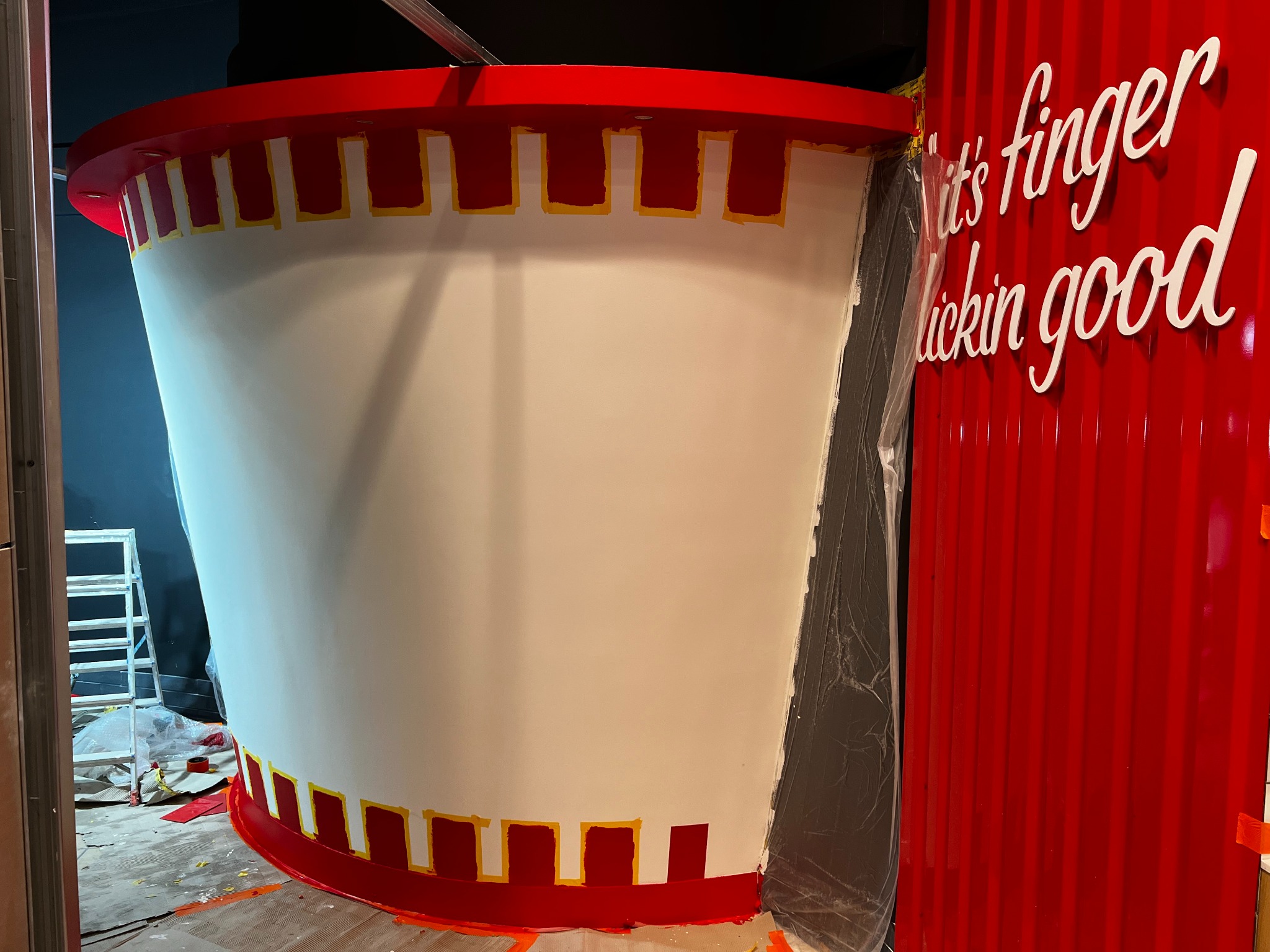 Wnętrze lokalu w trakcie malowania dekoracji imitującej kubełek KFC, z widocznymi taśmami malarskimi i napisem reklamowym na czerwonej ścianie.