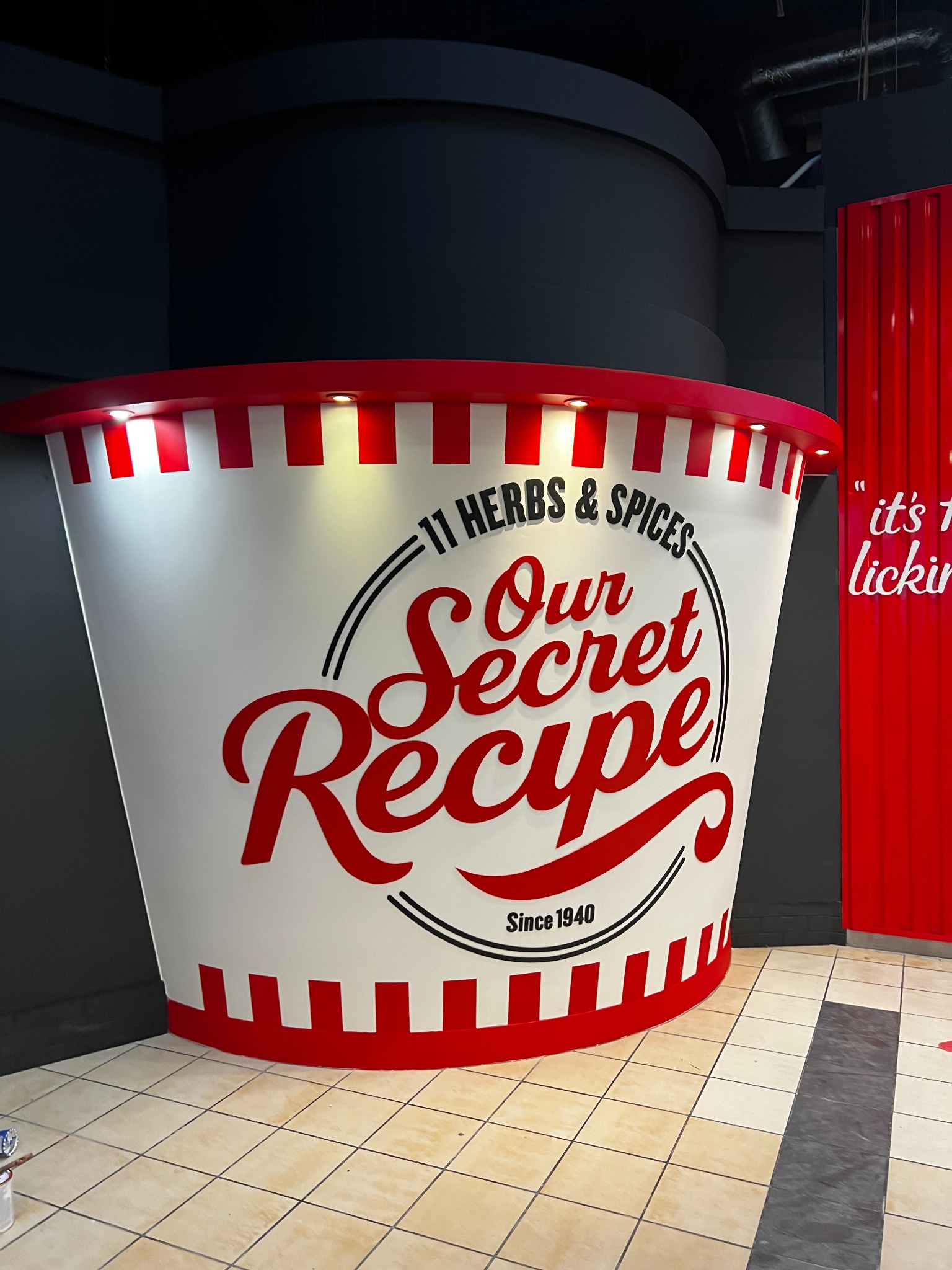 Wielkoformatowa reklama KFC w Warszawie: imitacja kubełka z napisem 'Our Secret Recipe' i czerwono-białymi paskami, umieszczona w lokalu.