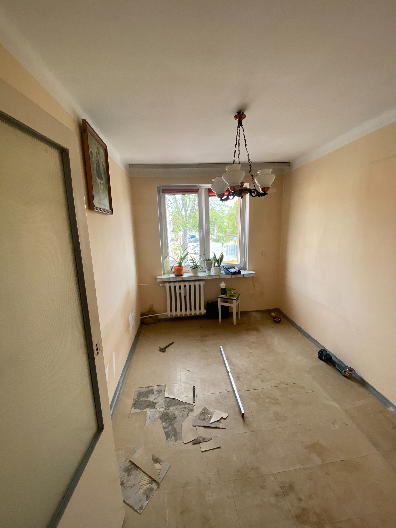 Pokój przed remontem: zerwane fragmenty podłogi, ściany w odcieniach beżu, okno z roślinami, wisząca lampa retro i narzędzia budowlane w rogu.