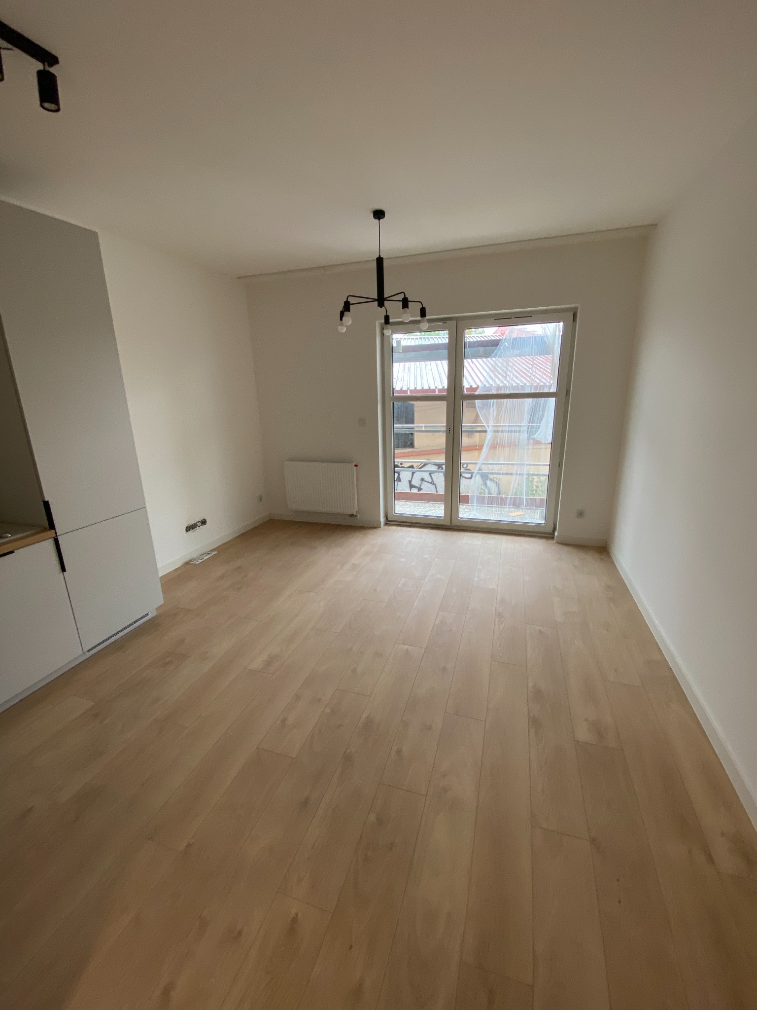 Puste, jasne pomieszczenie z drewnianą podłogą, białymi ścianami i balkonem. Widoczny grzejnik i minimalistyczna lampa sufitowa. Świeżo pomalowane ściany.