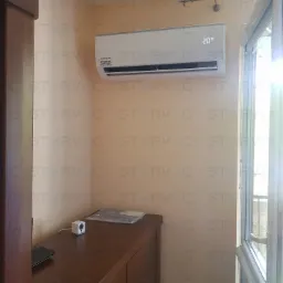 Biała klimatyzacja typu split marki Kaisai zamontowana na beżowej ścianie w pokoju, widoczny fragment okna balkonowego i ciemnobrązowa komoda z kamerą IP.