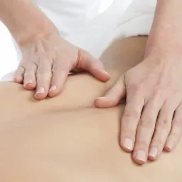 Dłonie masażysty uciskają plecy pacjenta, widoczne delikatne wgłębienia skóry pod naciskiem palców, białe prześcieradło w tle.