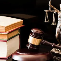 Brązowa statuetka Temidy z wagą i mieczem, drewniany młotek sędziowski na podstawce oraz trzy tomy kodeksów prawnych na czarnym, błyszczącym tle.