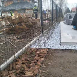 Montaż ogrodzenia panelowego z podmurówką, obok rabata z korą i młodymi sadzonkami, dalej białe kamienie dekoracyjne i płytki tarasowe, w tle dom w trakcie budowy i pracownicy.