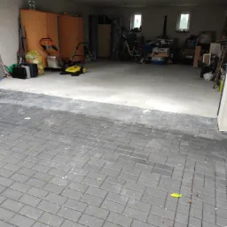 Wnętrze garażu z widocznymi szafkami, rowerem, kartonami i narzędziami, z kostką brukową przed wjazdem i pojedynczym żółtym liściem na niej.