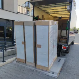 Dwa duże, zapakowane w folię meble, prawdopodobnie szafy, na platformie załadunkowej otwartego busa transportowego, na tle nowoczesnego budynku mieszkalnego z balkonami.