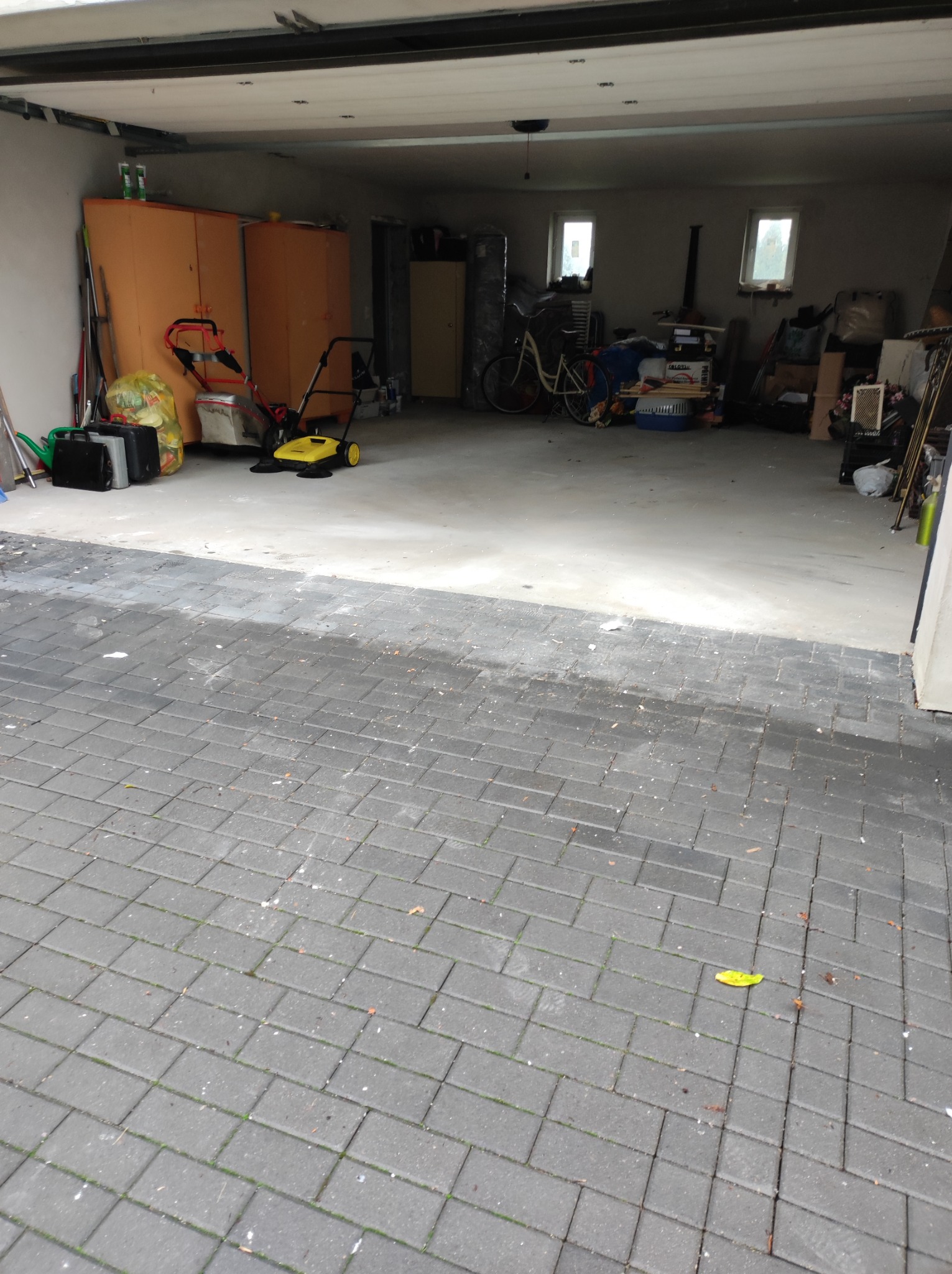 Wnętrze garażu z widocznymi szafkami, rowerem, kartonami i narzędziami, z kostką brukową przed wjazdem i pojedynczym żółtym liściem na niej.