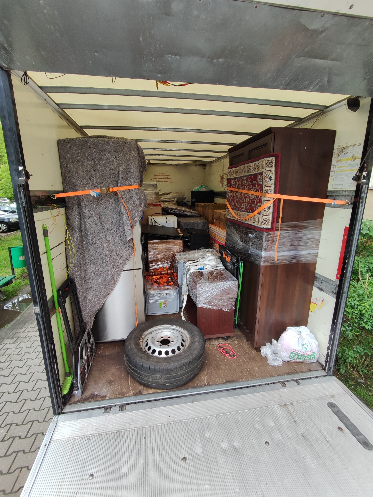 Wnętrze furgonetki załadowane meblami, sprzętem AGD i kartonami zabezpieczonymi folią stretch i pasami transportowymi podczas przeprowadzki. Widoczna opona samochodowa i akcesoria do sprzątania.