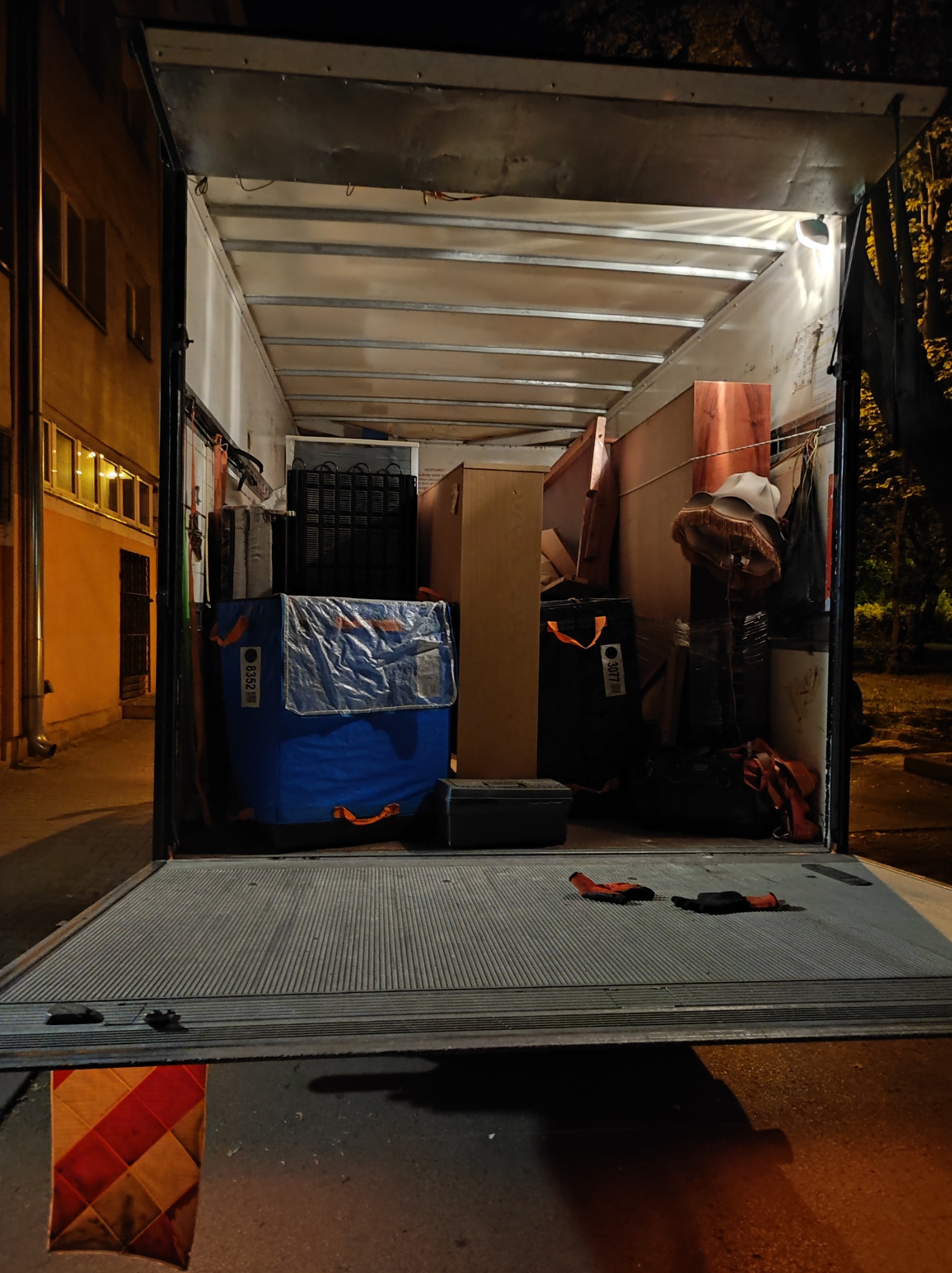 Wnętrze załadowanego busa z elementami wyposażenia domu, widoczne kartony, lampa z abażurem i niebieska torba transportowa z numerem 8352, na rampie leżą rękawice robocze.