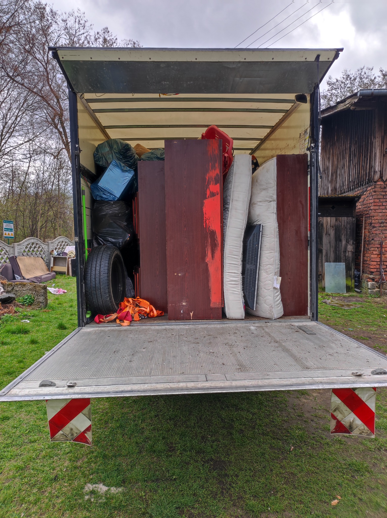 Otwarta paka samochodu dostawczego wypełniona meblami, materacami, oponą i workami z rzeczami, na zewnątrz widoczny dom i zieleń.