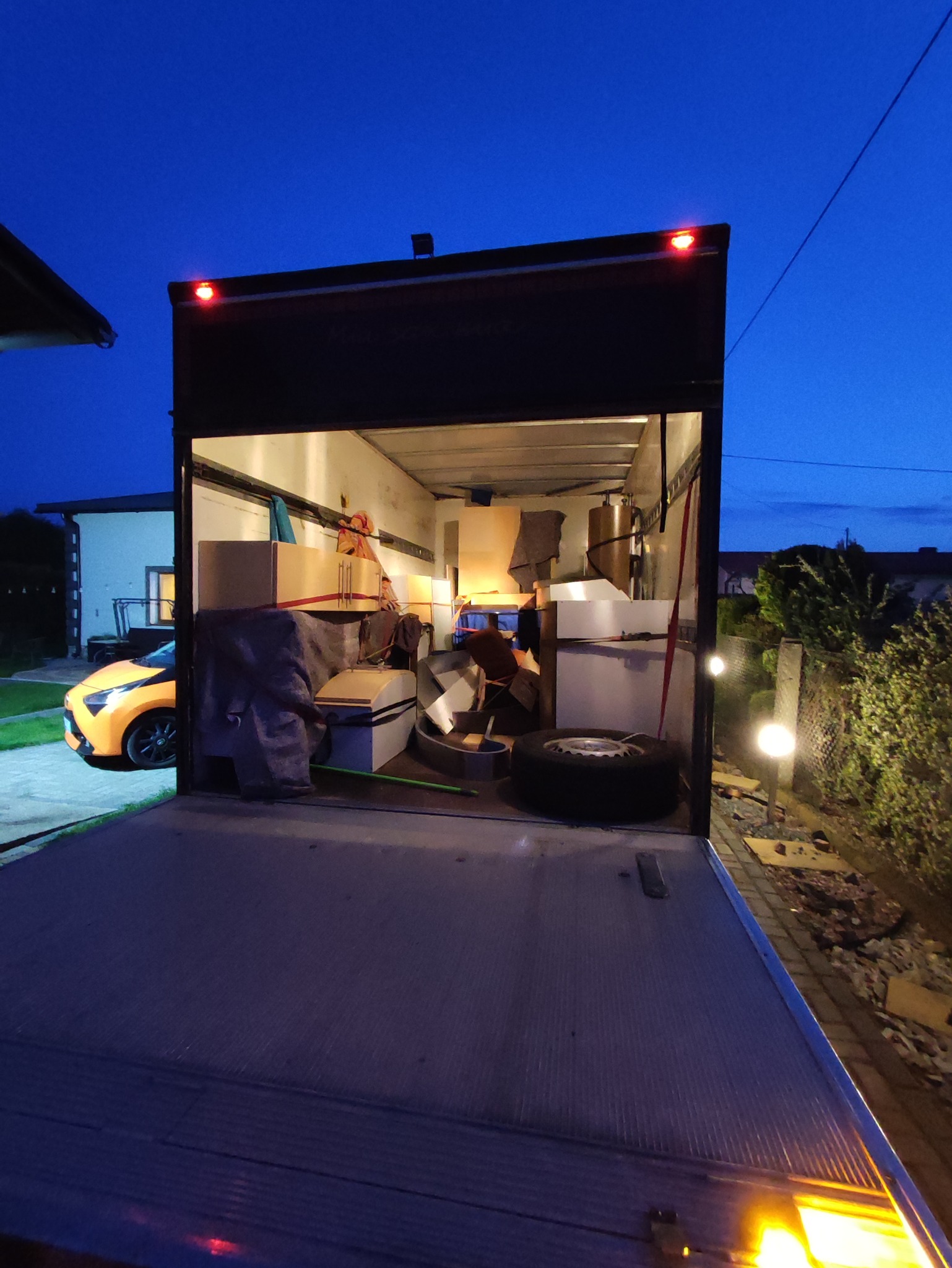 Wnętrze załadowanego busa meblowego z otwartą klapą, widoczne kartony, meble i opona samochodowa, w tle dom i pomarańczowy samochód w wieczornym oświetleniu.