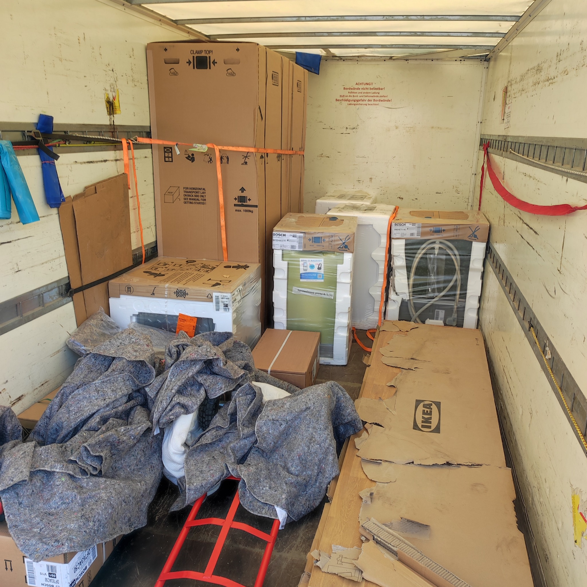 Wnętrze furgonetki wypełnionej kartonami z meblami i sprzętem AGD marki Bosch i IKEA, zabezpieczonymi pasami transportowymi i kocami ochronnymi, gotowe do transportu.