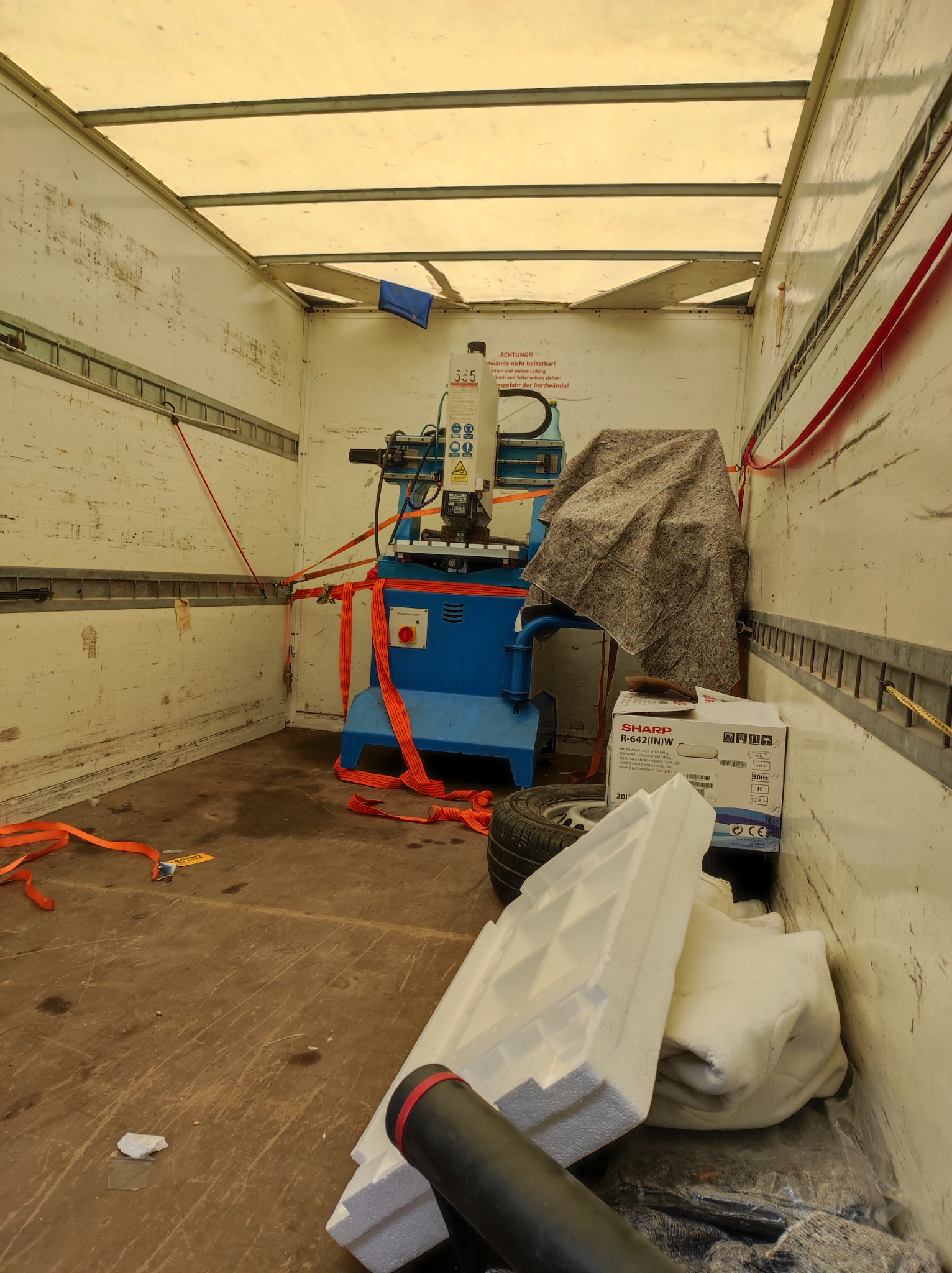 Wnętrze brudnej przestrzeni ładunkowej busa z zabezpieczoną maszyną do obróbki metalu, przykrytą szarą tkaniną, obok karton Sharp R-642(IN)W, opona, styropianowe elementy ochronne i czerwone pasy...