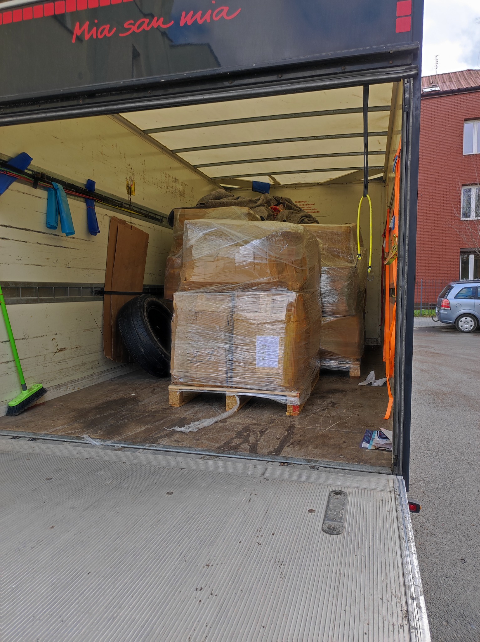 Wnętrze przestrzeni ładunkowej wypełnionej kartonami owiniętymi folią stretch na paletach, dodatkowo widoczna opona samochodowa i zielona zmiotka.