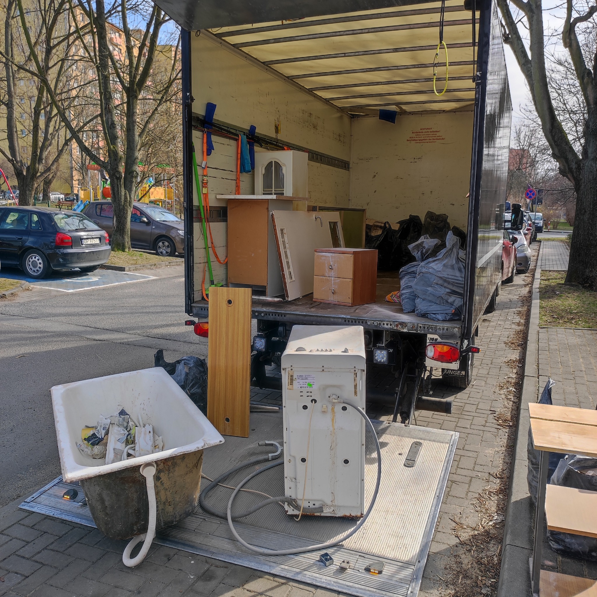 Opróżniana platforma załadunkowa ciężarówki z meblami, starą wanną i pralką, widok z tyłu na tle osiedla mieszkaniowego.