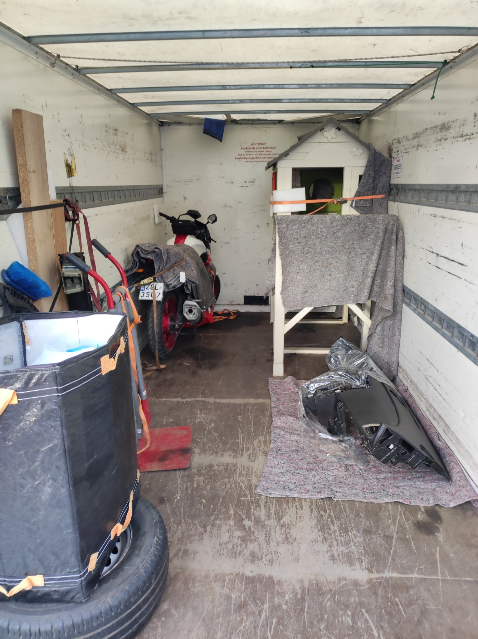 Wnętrze paki busa z załadowanym motocyklem przykrytym kocem, domkiem dla zwierząt zabezpieczonym pasami transportowymi, czarnym pojemnikiem na oponie, elementami karoserii samochodowej owiniętymi...