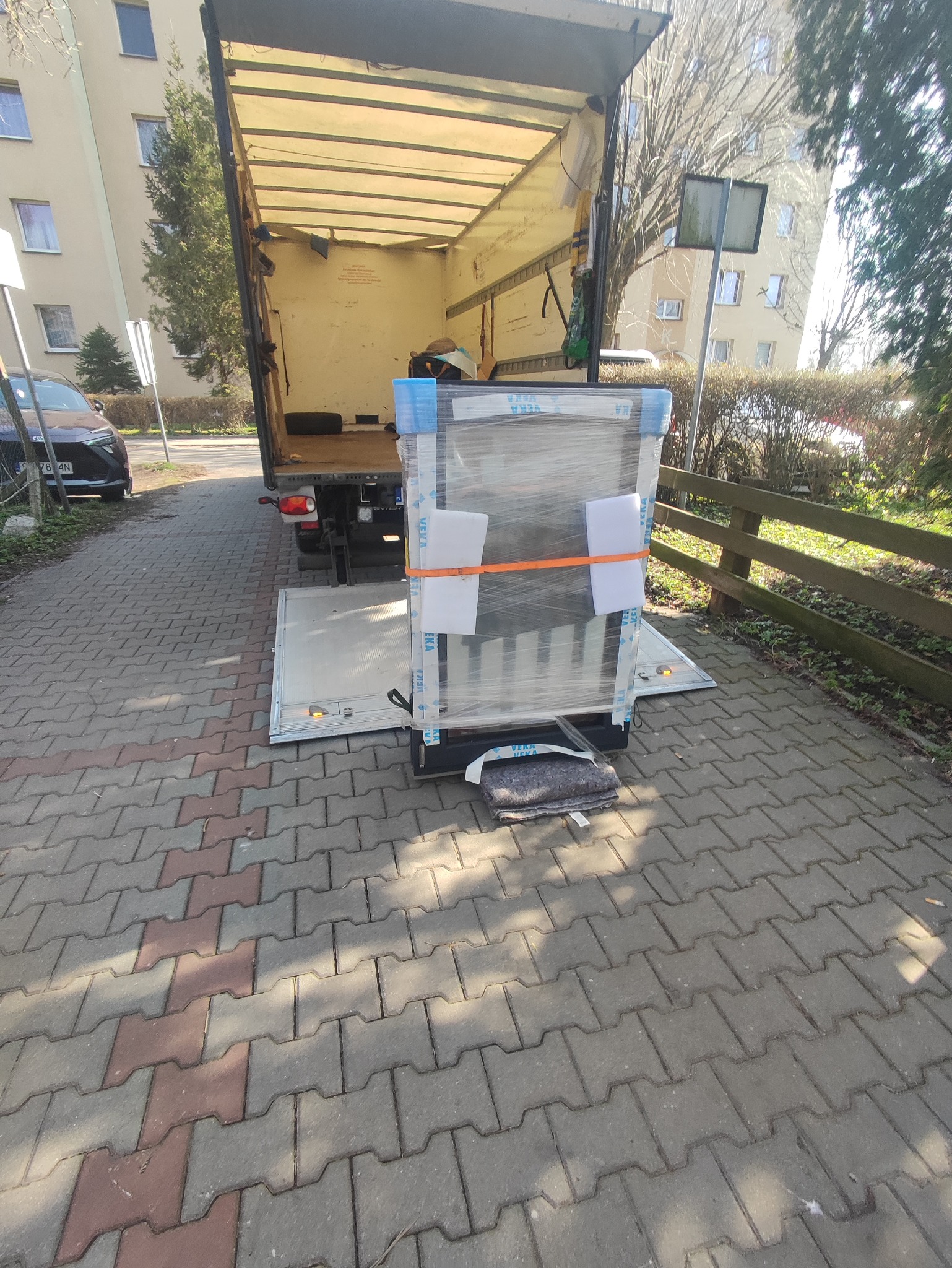 Okno owinięte folią i zabezpieczone pianką, na platformie windy samochodu dostawczego, z widocznym budynkiem mieszkalnym w tle.