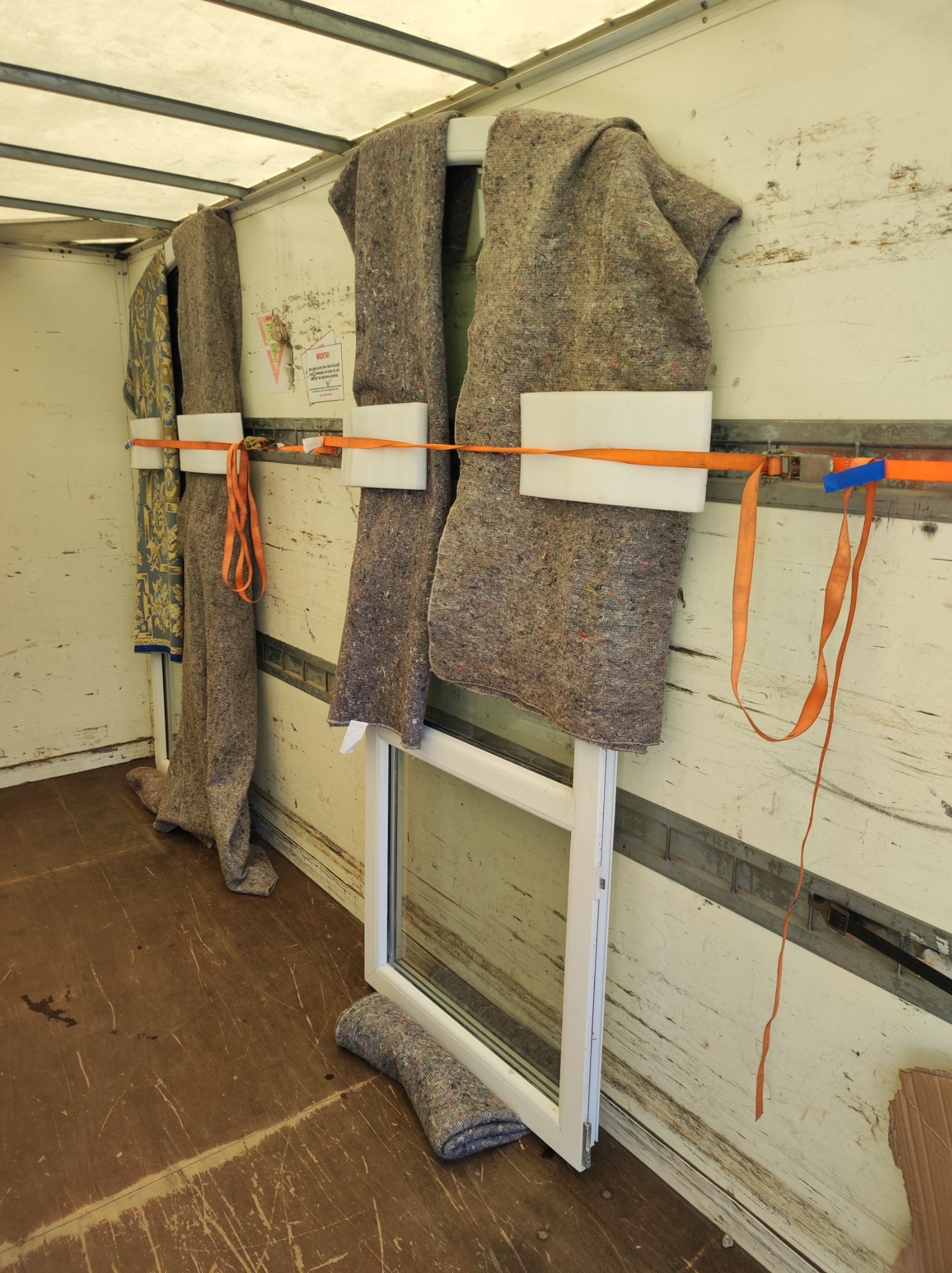 Wnętrze skrzyni ładunkowej z oknem PCV zabezpieczonym kocami transportowymi, przymocowanymi pomarańczowymi pasami transportowymi i białymi piankowymi przekładkami.