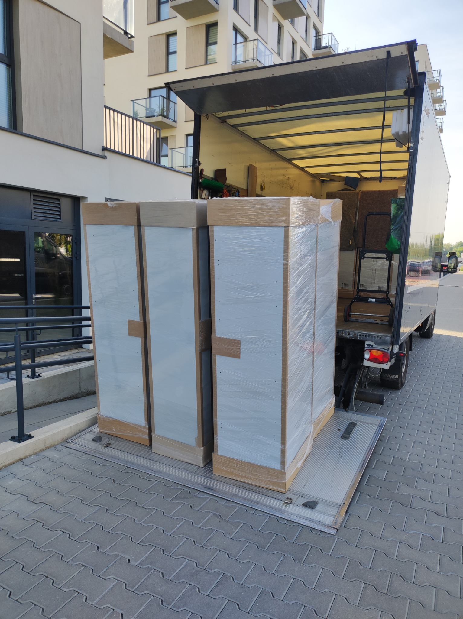 Dwa duże, zapakowane w folię meble, prawdopodobnie szafy, na platformie załadunkowej otwartego busa transportowego, na tle nowoczesnego budynku mieszkalnego z balkonami.
