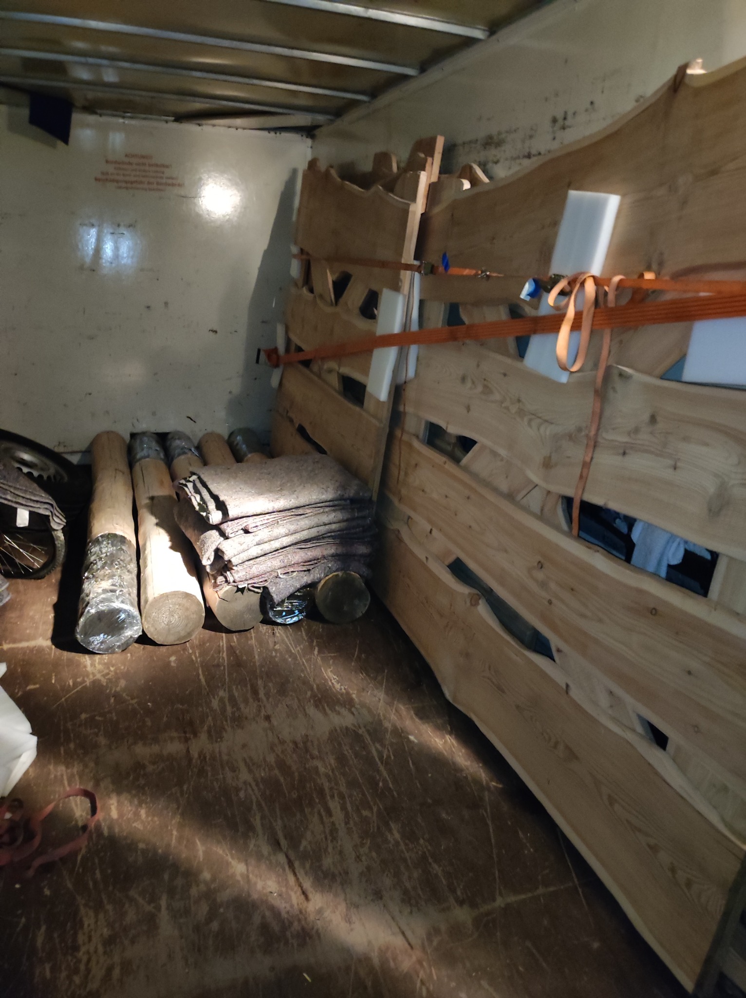 Wnętrze brudnej przestrzeni ładunkowej z drewnianymi elementami konstrukcyjnymi zabezpieczonymi pomarańczowymi pasami transportowymi, obok leżą owinięte folią drewniane słupki i sterta koców...