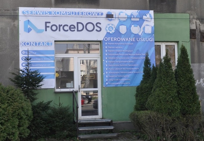 Fasada budynku firmy ForceDOS z szyldem oferowanych usług komputerowych, w tym naprawa komputerów, odzyskiwanie danych, montaż sieci i inne. Widoczne wejście do lokalu i zieleń przed budynkiem.
