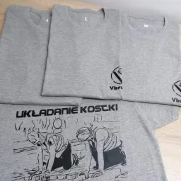 Stos szarych koszulek z nadrukiem: logo firmy i grafika przedstawiająca układanie kostki brukowej, różne rozmiary L i M.