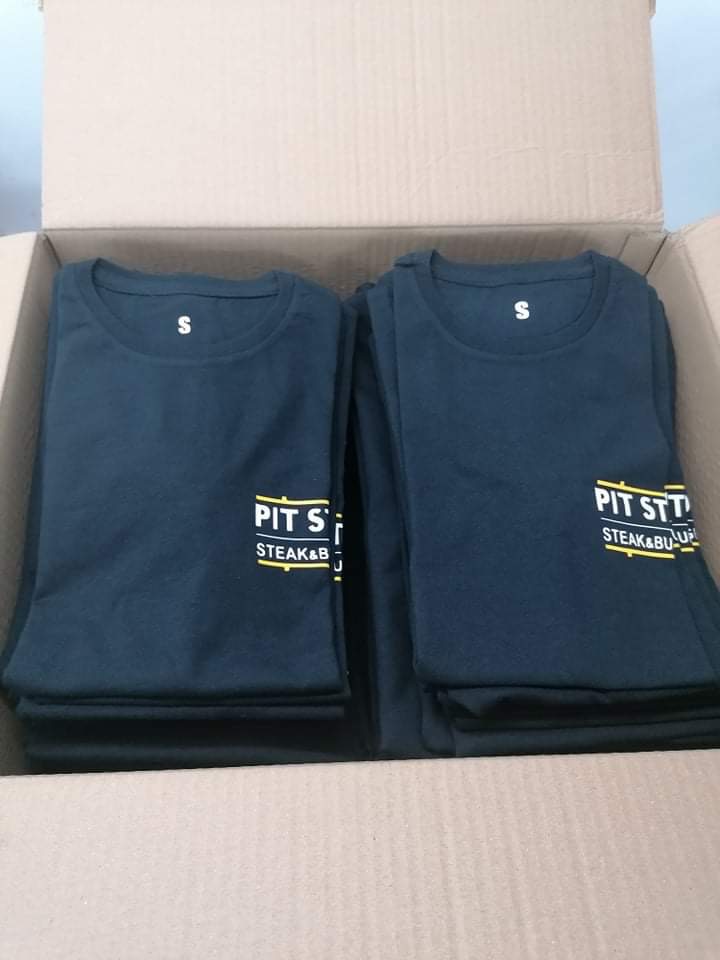 Stos czarnych koszulek z nadrukiem 'PIT STOP STEAK&BURGER' w kartonowym pudełku, wykonanych w Głownie.