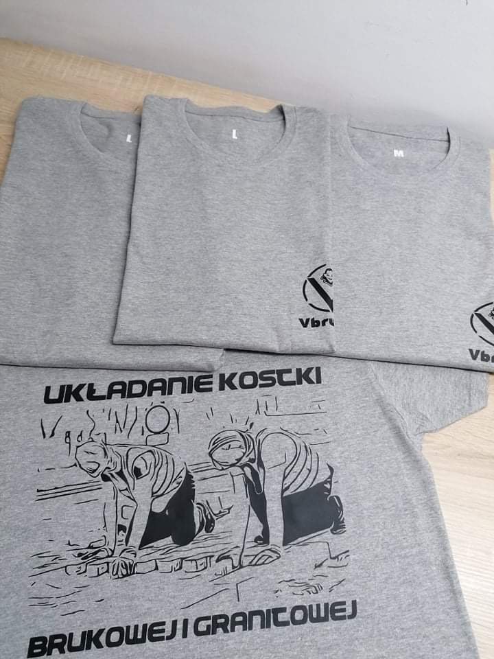 Stos szarych koszulek z nadrukiem: logo firmy i grafika przedstawiająca układanie kostki brukowej, różne rozmiary L i M.