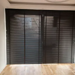 Ciemne żaluzje drewniane zamontowane w oknie, z widocznymi promieniami słońca wpadającymi do pomieszczenia, oświetlając parkiet