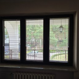 Mariusz - Okno z trzema skrzydłami wyposażone w poziome żaluzje drewniane, przez które widać zieleń ogrodu i elementy architektury budynku.