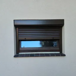 Okna z częściowo opuszczoną, brązową roletą zewnętrzną, widoczne odbicie nieba i drzew w szybie, jasna elewacja budynku.