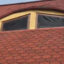 Dwa okna dachowe z moskitierami w żółtej ścianie na tle czerwonego dachu z gontu bitumicznego. Ujęcie z dołu, widok na zamontowane siatki.