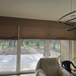 Mariusz - Beżowe, zwinięte  w górnej części okna, materiałowe osłony przeciwsłoneczne w salonie z widokiem na las. Nowoczesna lampa i zegar na ścianie.