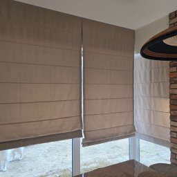 Mariusz - Beżowe rolety rzymskie zamontowane w oknach, widoczne z wnętrza pomieszczenia z ceglaną ścianą i dekoracyjną lampą. Na zewnątrz widać psa.