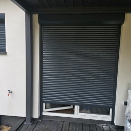 Mariusz - Grafitowa roleta zewnętrzna częściowo opuszczona na oknie domu z jasną elewacją. Widoczny fragment otwartego okna i worek z odpadami po montażu.