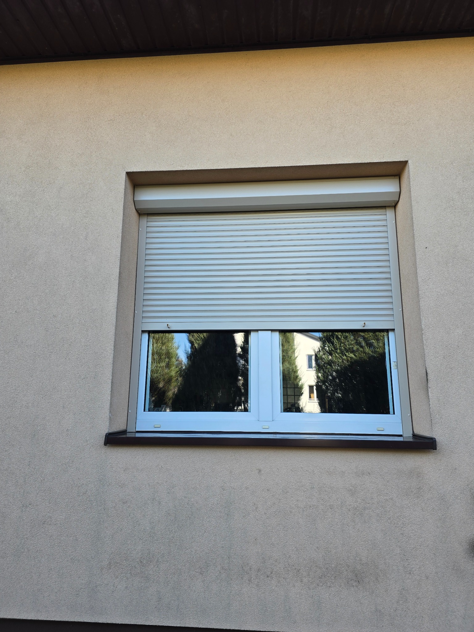 Zamknięta, kremowa roleta zewnętrzna na oknie z białymi ramami; w szybie odbijają się drzewa i budynek.