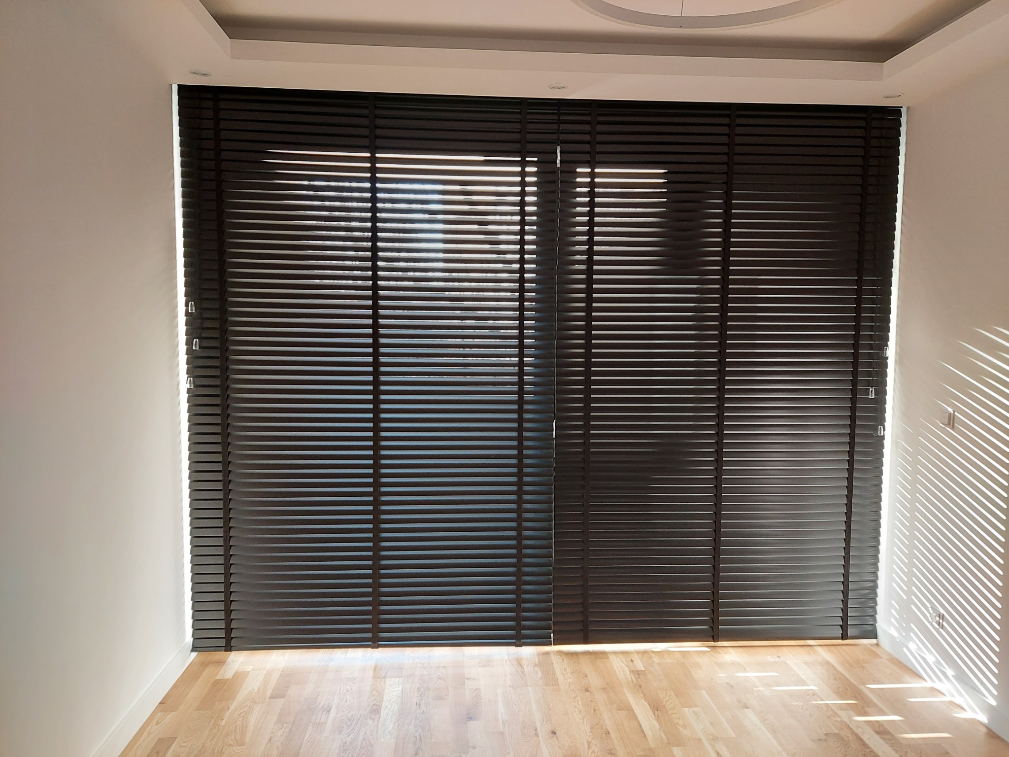 Ciemne żaluzje drewniane zamontowane w oknie, z widocznymi promieniami słońca wpadającymi do pomieszczenia, oświetlając parkiet