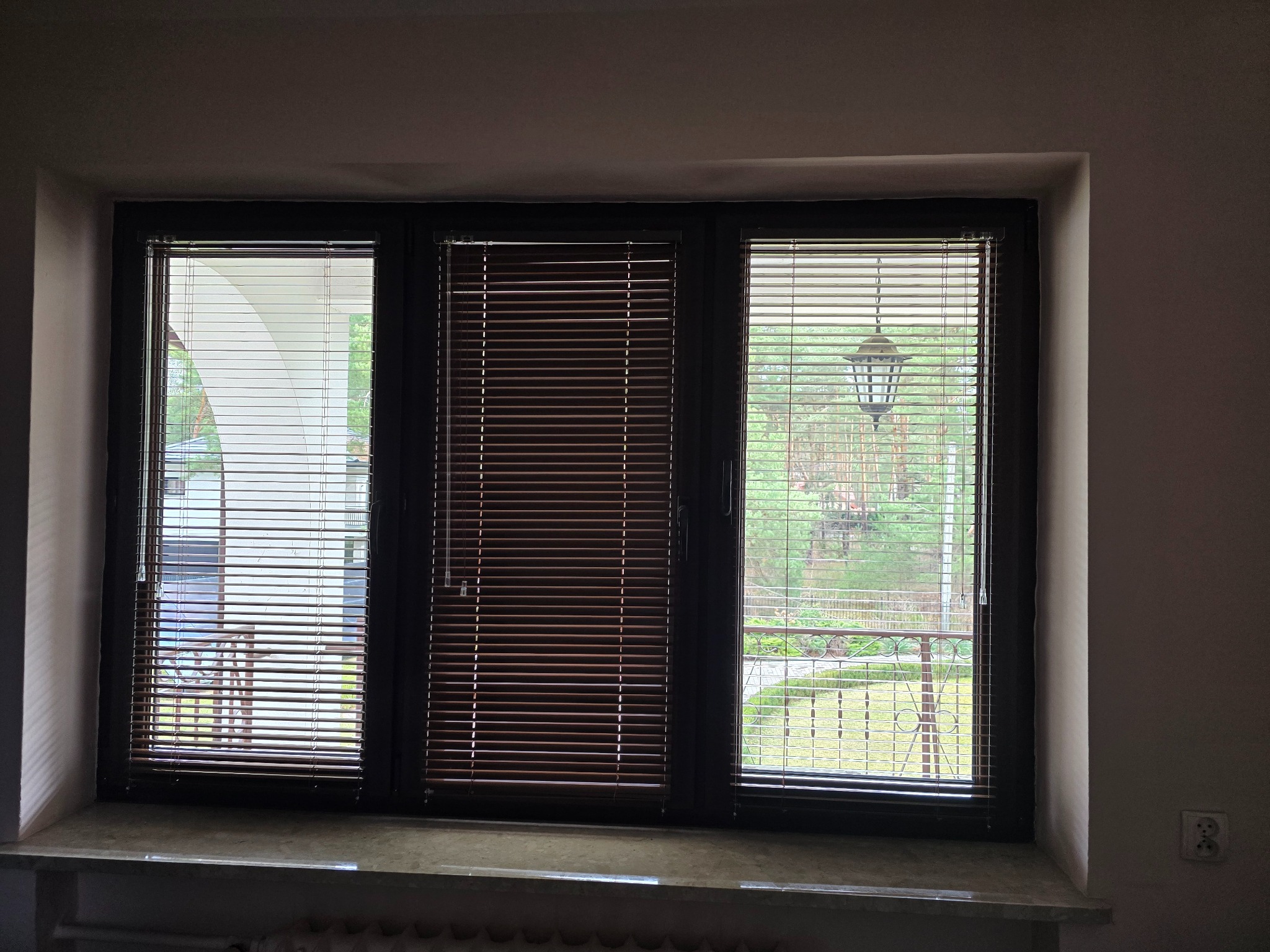 Trzyczęściowe okno z zamontowanymi żaluzjami: białe po lewej, drewniane pośrodku i białe po prawej, widok na zewnątrz z elementami architektury i zieleni.