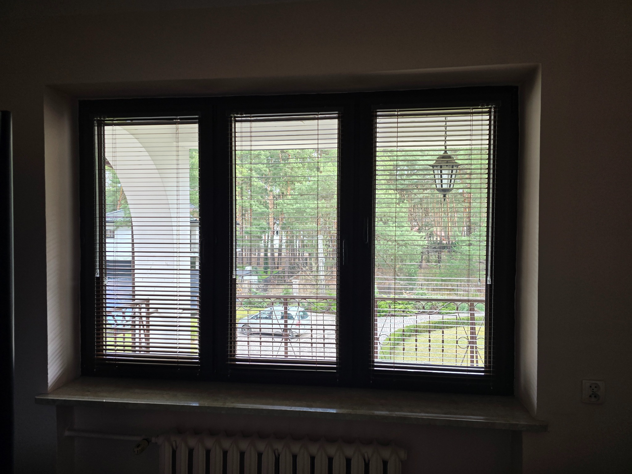 Okno z trzema skrzydłami wyposażone w poziome żaluzje drewniane, przez które widać zieleń ogrodu i elementy architektury budynku.