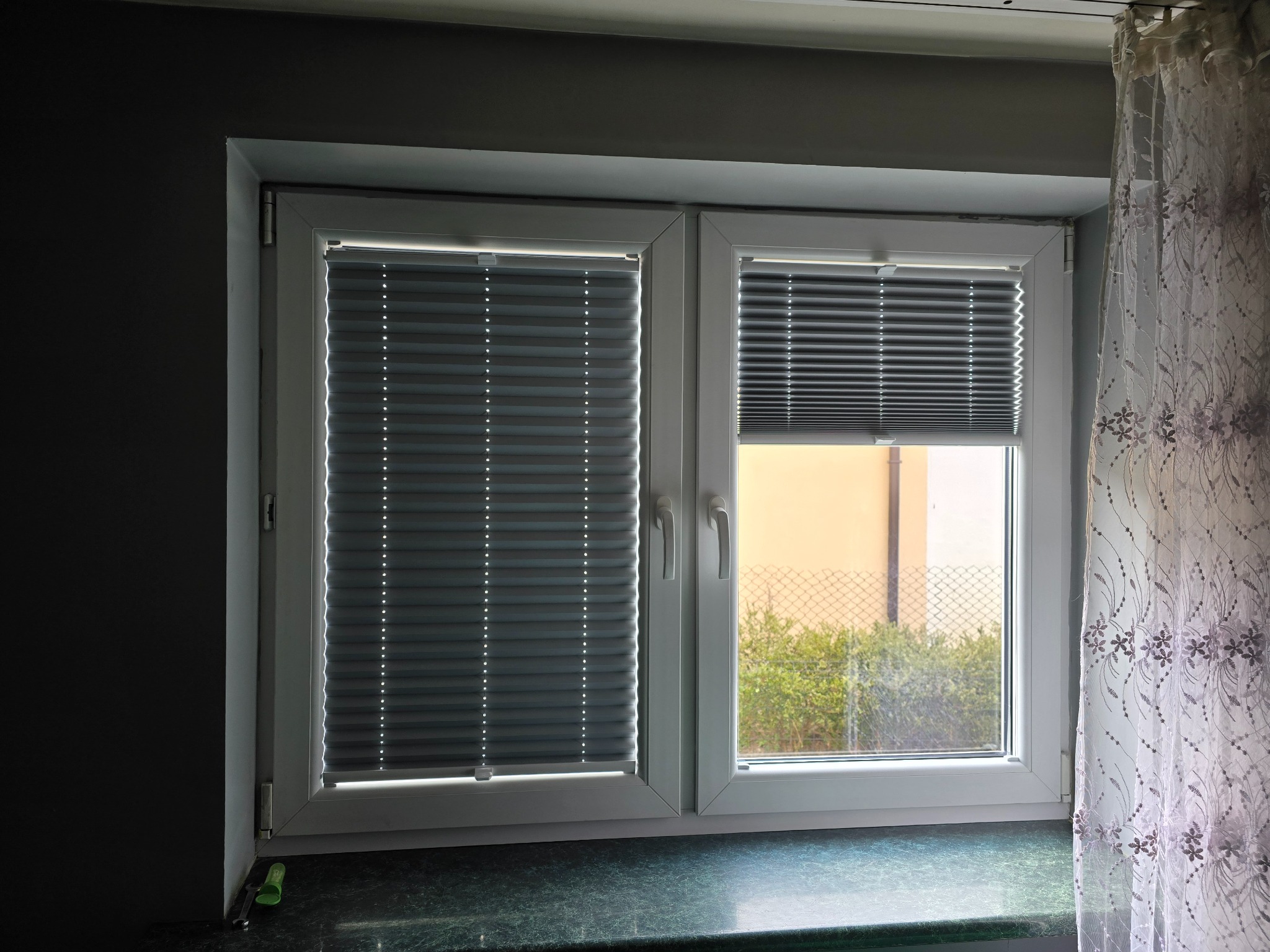 Okno PCV z szarymi plisami, jedna opuszczona całkowicie, druga częściowo, widok na zewnątrz przez siatkę ogrodzeniową i zieleń, fragment firanki z kwiatowym wzorem po prawej.