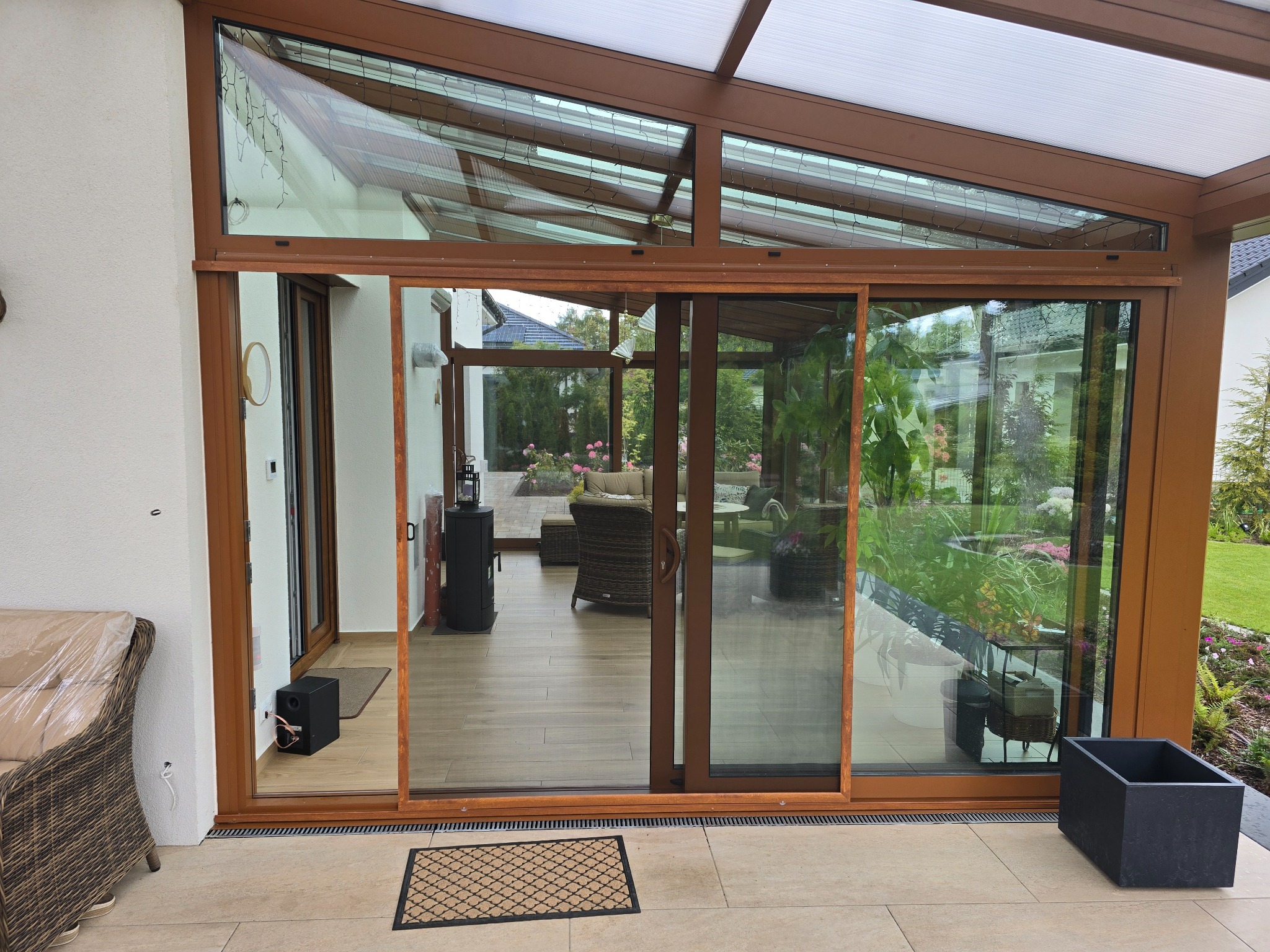 Zadaszony taras z przesuwnymi drzwiami balkonowymi z brązowymi ramami. Widok na ogród i wnętrze domu. Mata podłogowa przed wejściem.