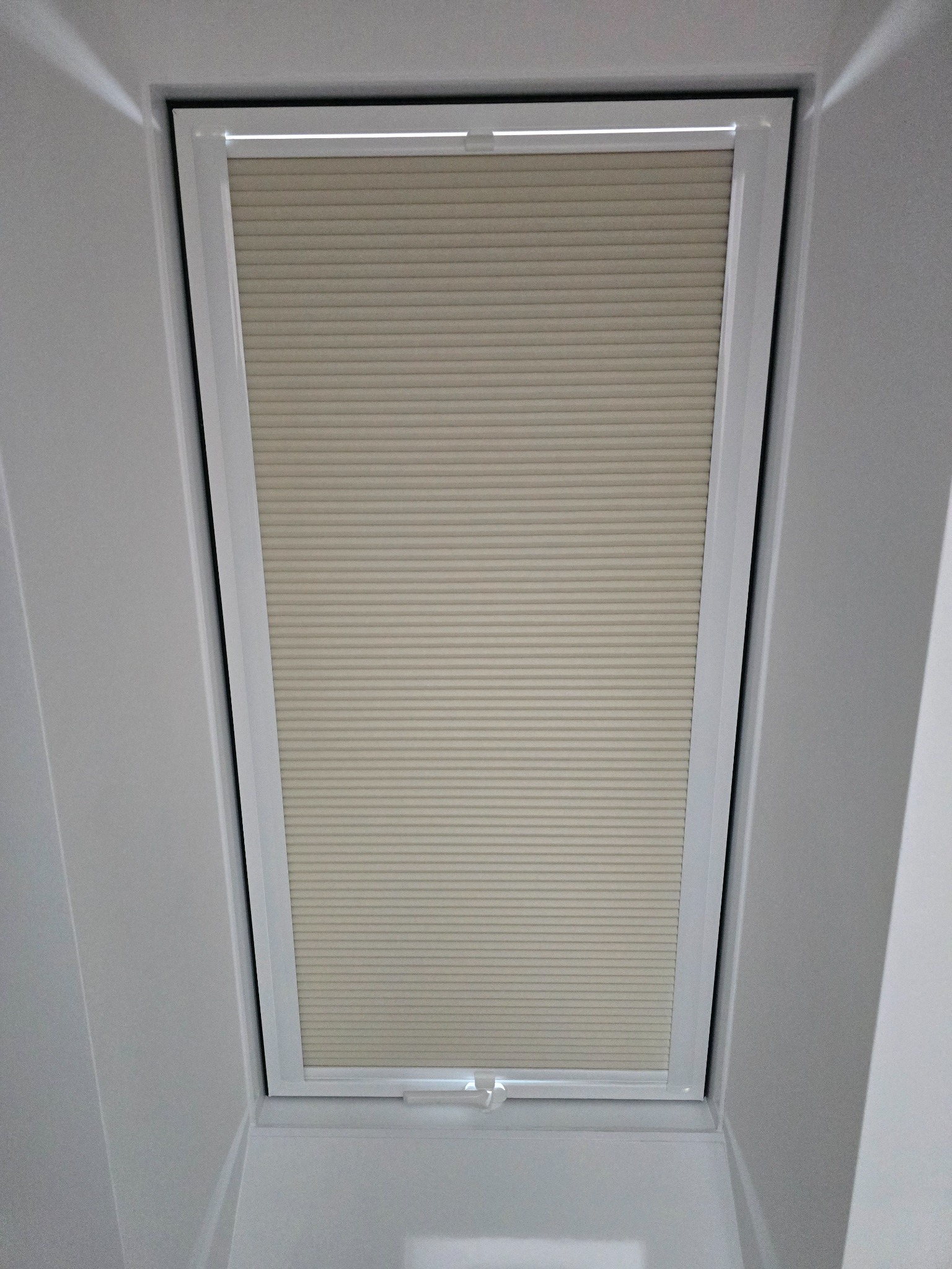 Zamknięta plisa okienna w białej ramie, zamontowana w oknie dachowym. Widok od dołu, prosto na okno. Jasne, neutralne kolory.