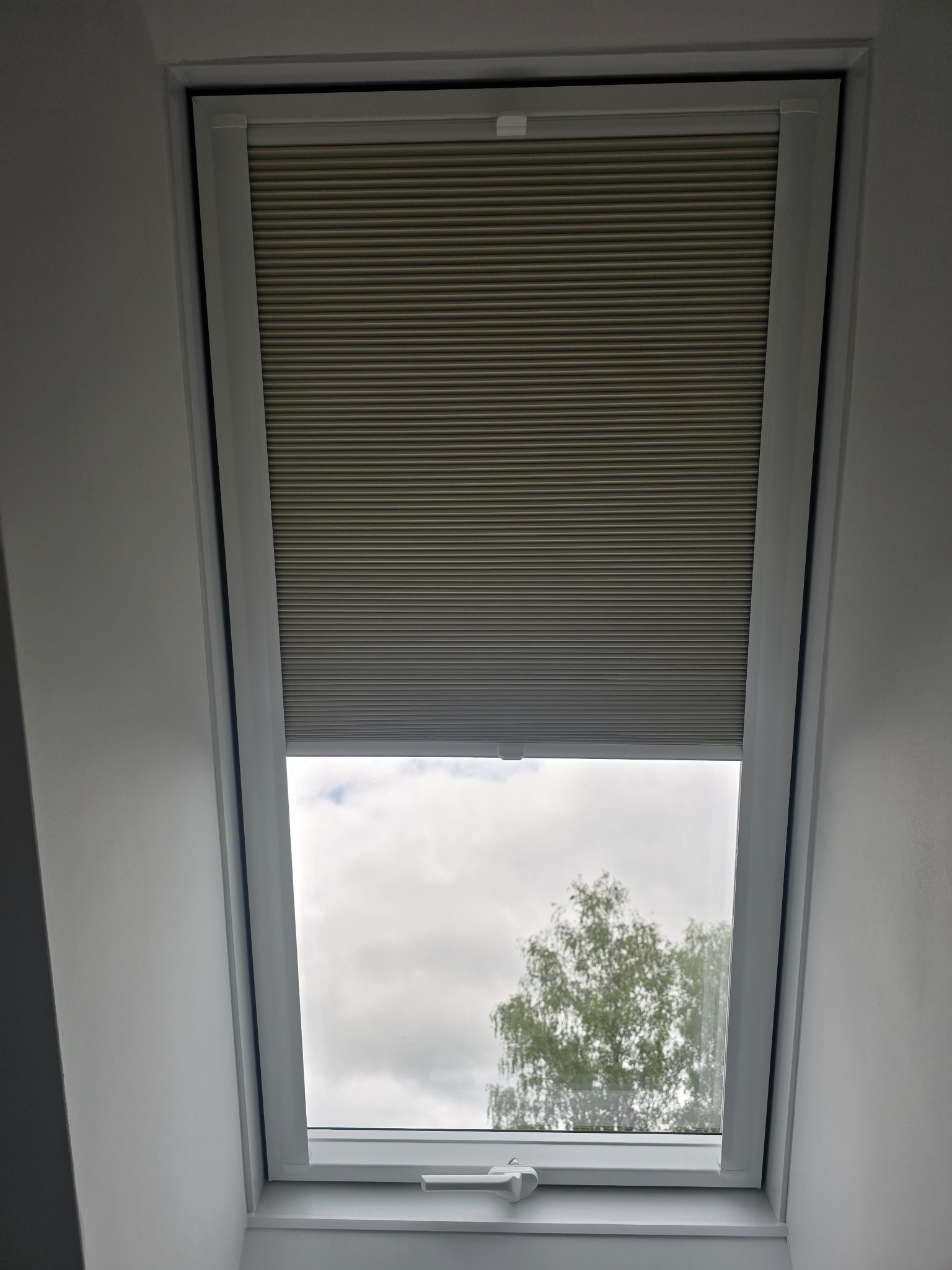 Szara plisa okienna zamontowana w oknie dachowym, widok na pochmurne niebo i drzewo. Nowoczesny design, stonowana kolorystyka, minimalistyczny styl.