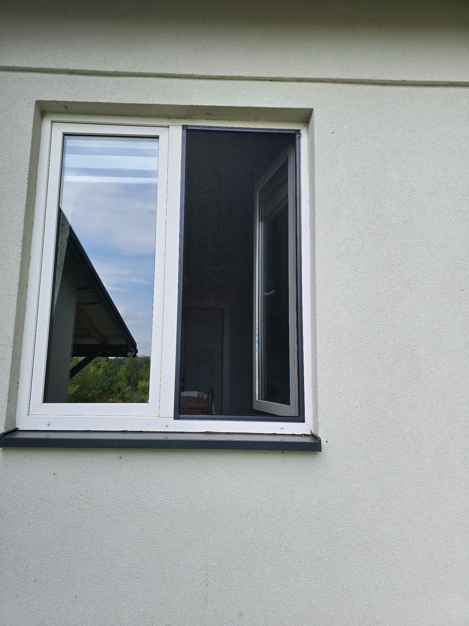 Białe okno z otwartą moskitierą na tle jasnoszarej, teksturowanej ściany budynku. Widoczne odbicie nieba i zieleni w szybie okna.