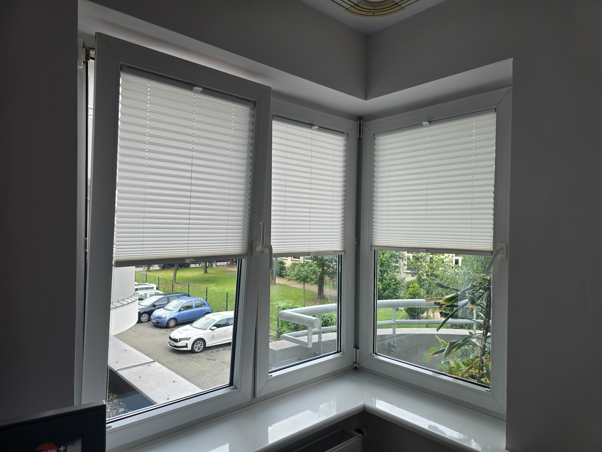 Okna z białymi plisami w kształcie trapezu, widok na parking i zieleń. Plisy częściowo zasłonięte, wpadające światło dzienne. Minimalistyczny design.