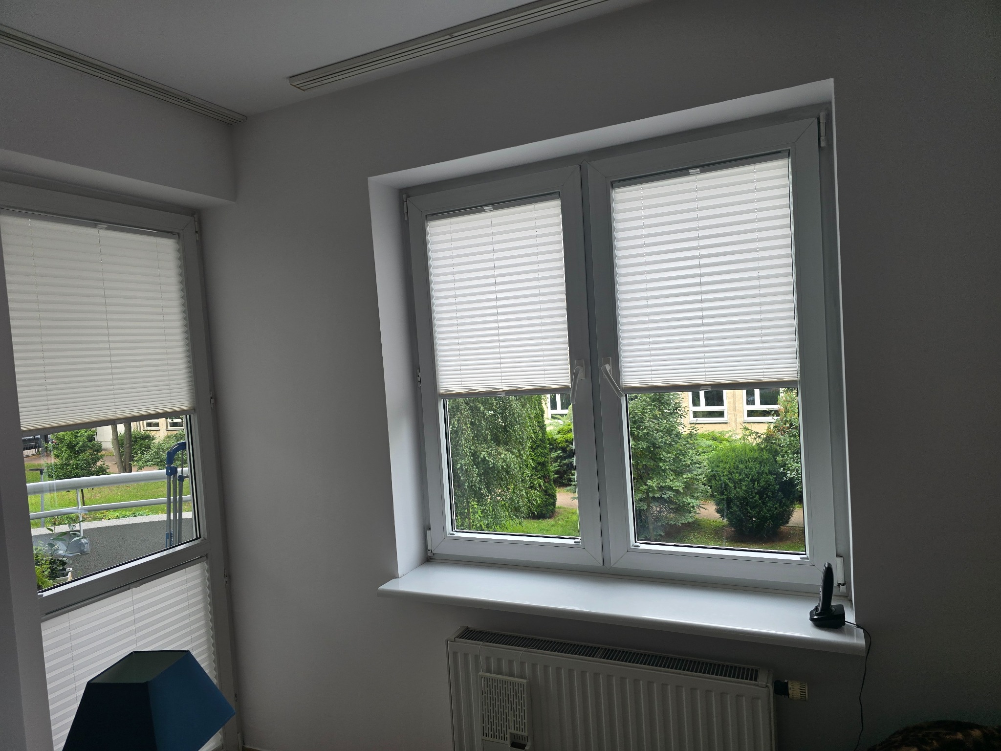 Białe, plisowane rolety w oknach z widokiem na zieleń. Rolety częściowo zasłonięte, widoczny grzejnik pod oknem i fragment balkonu z roślinami w drugim oknie.